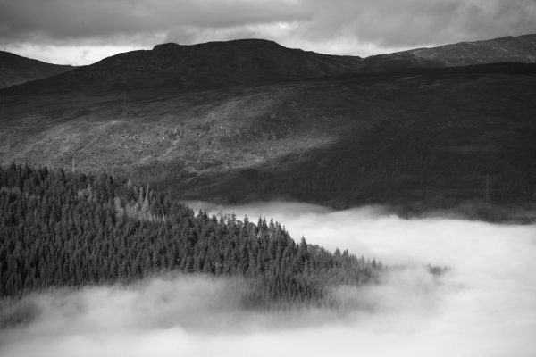 A black and white photograph of hills towering over a cloud inversion that looks like a floating white lake. The clouds are minging with the trees in a forest. 