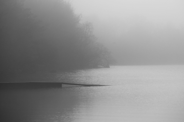 A black and white photograph of a muted and misty loch scene where a small jetty extends onto a clam water. 