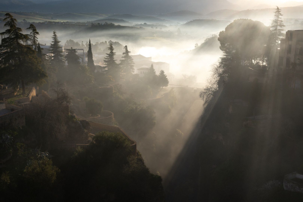 The view from the Puente Nuevo in Ronda in the morning mist. You can see into a gorge, with an old bridge in the background. There are also terraces with gardens and trees on both sides, as well as a few houses. In the background, you can see some hills, all backlit by the sun.