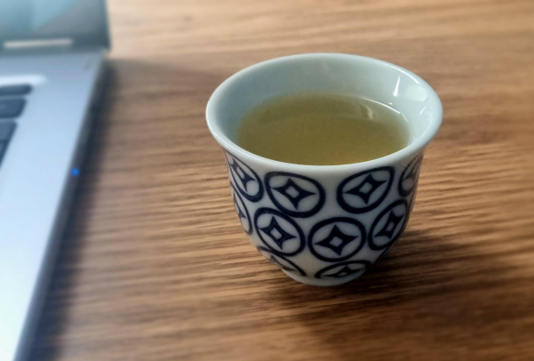 Small Japanese style tea cup next to a laptop. The cup is white with blue circles and inside each circle is a four pointed star shape. Inside the cup is a light green coloured liquid which is stewed lemon verbena 