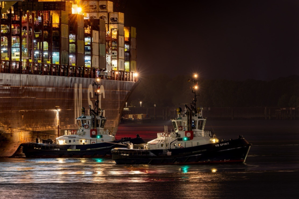 Zwei Schlepper unterstützen nachts ein großes Frachtschiff, das von Lichtern des Schiffes und der Umgebung beleuchtet wird. Das Frachtschiff hat bunte Container auf seinem Deck gestapelt, während die Schlepper aktiv im Wasser manövrieren.