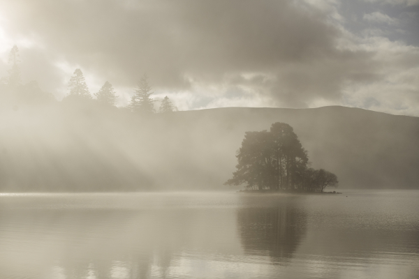 A colour photograph of mist over a Loch (actually a cloud inversion). There are sun rays coming through thr mist and a small island of trees in the still water.