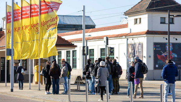 Außenaufnahme von Gebäuden der Messe Dresden. Im Bild sind zahlreiche Menschen abgebildet, die sich vor den Gebäuden finden. Fahnenmasten verweisen auf die Messe KarriereStart.