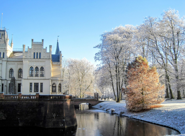 Bild einer winterlichen Szene: auf der linken Seite ist ein Teil eines neugotischen, kleinen Schlosses zu sehen, das von einer Mauer mit Wassergraben davor umgeben wird. Auf der rechten Bildseite ist der englische Park zu sehen mit hohen, mit Rauhreif bedeckten Bäumen. Der Himmel ist hellblau und klar.