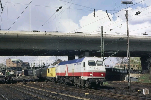 Eine Lokomotive der Baureihe 240 (TAURUS), lackiert in einem speziellen Design mit der Aufschrift "MAX 1074", zieht einen Zug, der aus verschiedenen Waggons, darunter einem gelben Wartungswagen, besteht. Die Szene spielt unterhalb einer großen Betonbrücke mit Oberleitungen und zahlreichen Gleisen im Vordergrund. Im Hintergrund sind Bahnhofsgebäude und städtische Strukturen zu erkennen. Die Umgebung deutet auf ein urbanes Bahnareal hin, wahrscheinlich ein wichtiger Bahnhof oder Rangierbahnhof.

