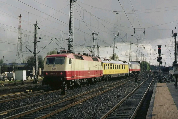 Eine Lokomotive der Baureihe 103 in beige-roter Lackierung, mit der Betriebsnummer 752 031-2, zieht einen Zug bestehend aus einem gelben Messwagen und einer weiteren Lokomotive der zuvor genannten Baureihe in beige-rot. Im Hintergrund ist eine weitere Lokomotive zu sehen, wahrscheinlich die Baureihe 240. Die Szene zeigt ein weitläufiges Gleisfeld mit Oberleitungen, Signaltechnik und einer typischen Bahninfrastruktur. Rechts im Bild befindet sich ein Bahnsteig mit einem Signal auf „Halt“. Der Himmel ist bewölkt, mit einigen Sonnenstrahlen, die die Szenerie beleuchten.

