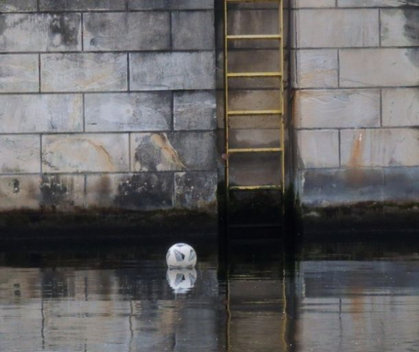 auf einem Fluss schwimmt ein weißer Ball mit grauen Mustern, im Hintergrund ist eine Wand aus rechteckigen Steinen zu sehen, an welcher eine gelbe Leiter angebracht ist. Der Ball ist bereits kurz vor der Leiter und bewegt sich von links nach rechts.