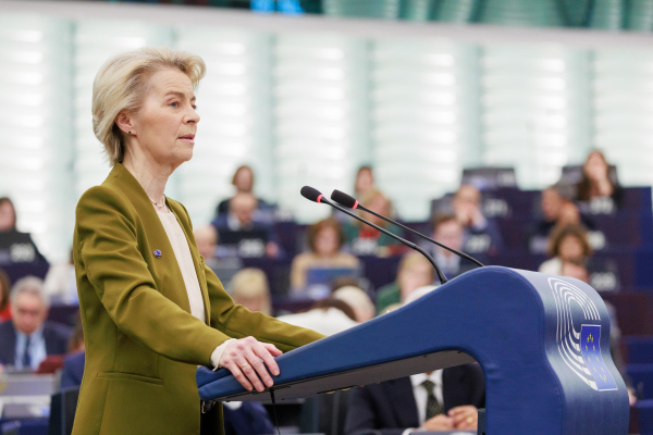 President von der Leyen delivering a speech at a podium in a formal setting, with an audience seated in the background. President wears a green blazer and stands confidently, addressing the assembly. 