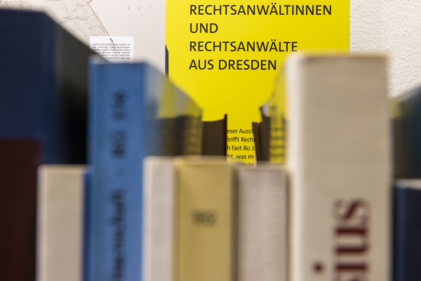 Nahaufnahme eines Bücherregals. Man sieht man Vordergrund Buchrücken. Im Hintergrund eine Tafel der Ausstellung. Sie ist gelb und zeigt den Text "Rechtsanwältinnen und Rechtsanwälte aus Dresden"