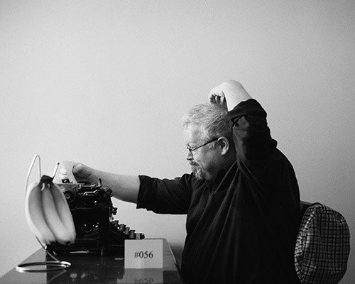 Mark A. Rayner, scratching his head as he looks at paper in his typewriter. there is a stand of bananas next to it and a sign that says #056