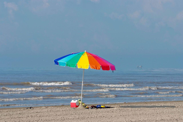 Das Foto zeigt einen regenbogenfarbigen Sonnenschirm am Strand. Im Hintergrund ist das Meer zu sehen