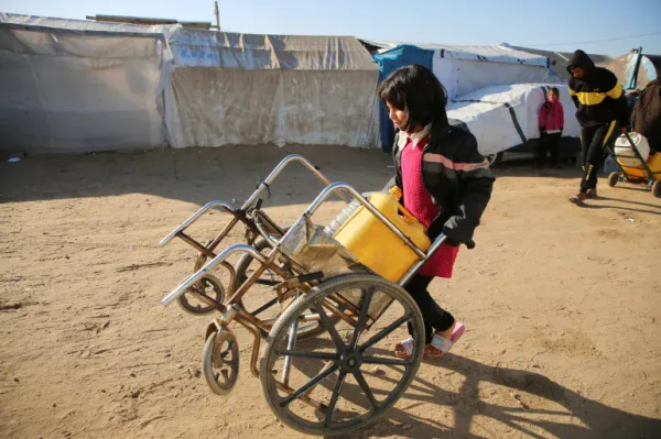 A girl pushing a wheelchair with a water container, Hatem Khaled