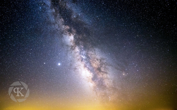 The core of the Milky Way arcs vertically into the sky. The orange-tinted glow of city lights along the horizon fade in a star-filled tapestry into the inky black void above.