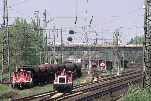 Ein Güterbahnhof mit mehreren Gleisen, auf denen rote Rangierlokomotiven verschiedene Güterwagen bewegen. Im Vordergrund stehen zwei Rangierloks neben beladenen Schüttgutwagen. Im Hintergrund sind weitere Gleise und Güterzüge zu sehen. Oberleitungen und Masten durchziehen das Bild und verdeutlichen die komplexe Infrastruktur des Bahnhofs. Im oberen Bildbereich führt eine Straßenbrücke über die Gleisanlagen, dahinter sind Bäume und ein heller Himmel erkennbar. Die Szene wirkt geschäftig und industriell.

