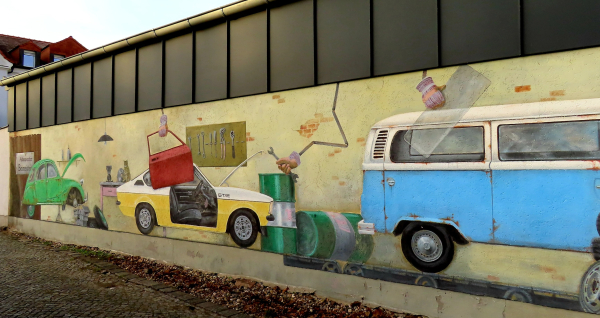 Fassade einer Autowerkstatt. 

Grünes, gelbweißes Auto, VW Bulli in blauweiß.
Die Autos defekt.
Geöffnete Motorhaube.
Entfernte Tür beim nächsten Auto, die abgehoben wird.

Ölfässer liegen rum.
Werkzeugtasche an der Wand.

Im Prinzip das Innere der Werkstatt auf die Fassade gemalt