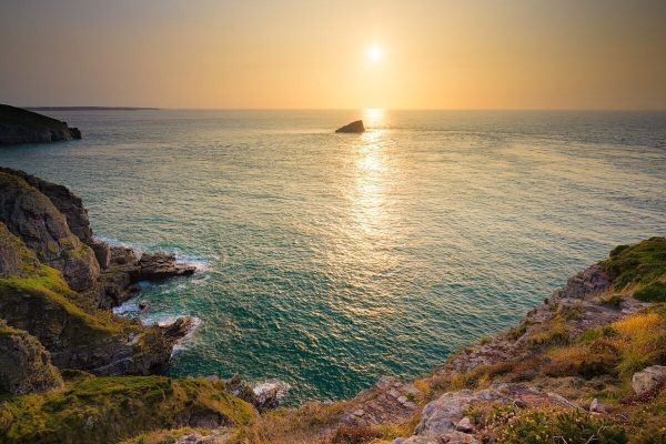 Zu sehen ist ein Sonnenuntergang über dem Meer vor einer Felsküste.