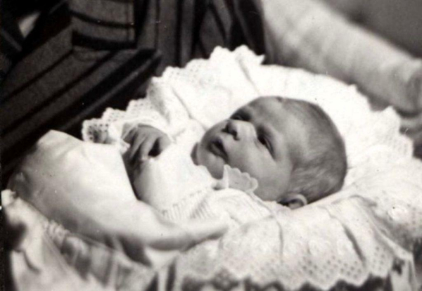 A vintage black-and-white photo of a newborn in a lace-trimmed outfit lying in a bassinet.