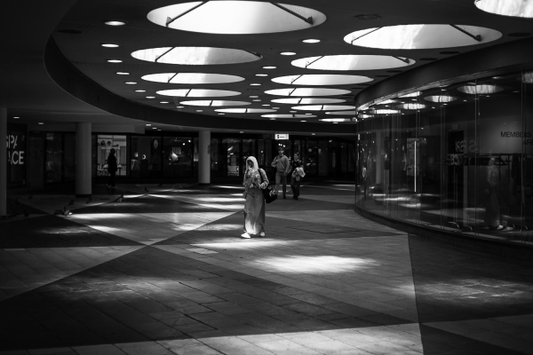 People walking through a shopping arcade. Spotlights fall through round openings in the roof. 