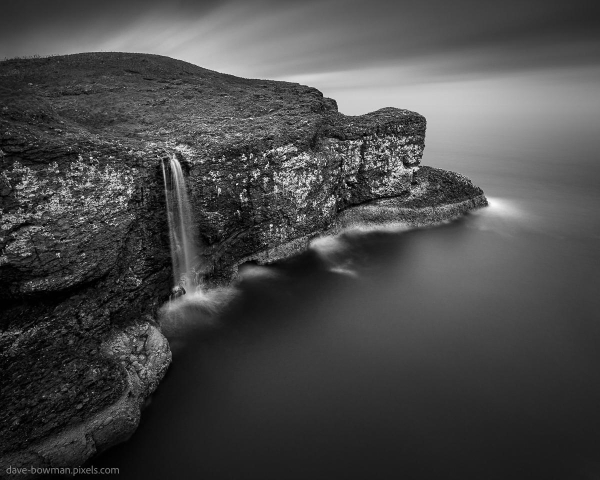 A photograph of a waterfall cascading over rugged cliffs at Crawton in Aberdeenshire, Scotland. The flowing water appears silky and smooth due to the long exposure, contrasting with the jagged texture of the rock face. The monochrome tones highlight the dramatic interplay of light and shadow, capturing the raw beauty of the Scottish coastline.