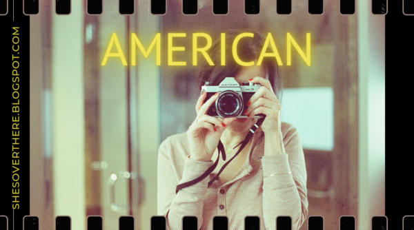 Screenshot from Youtube video "AMERICAN" in yellow with dark haired woman taking a self with 35mm camera. Sprockets!