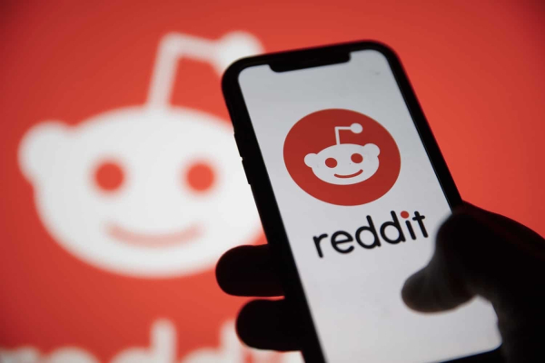 Eine Hand bedient ein iPhone, auf dem das Reddit-Logo und -Schriftzug zu sehen sind. Im Hintergrund sieht man unscharf ein großes weißes Reddit-Logo, das von orangeroter Farbe umgeben ist. 