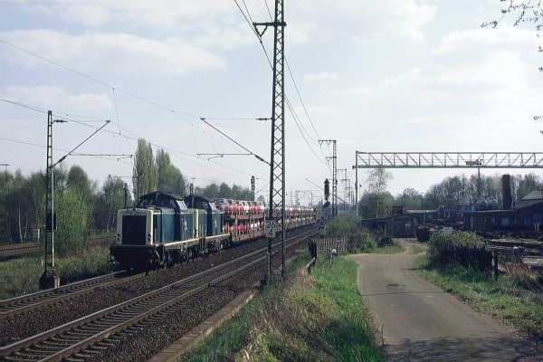 Eine blaue Lok tuckert über parallele Gleise, hinten dran ein paar rote Waggons. Das Ganze spielt in einer ländlichen Gegend, mit Stromleitungen oben drüber und ein paar Bäumen und Gebäuden in der Ferne. Rechts parallel zu den Gleisen verläuft ein breitgefahrener Feldweg, welcher zu einem Gewerbehof mit mehreren Schuppen und Industriehallen führt. Über der Einfahrt hängt eine sehr hohe Truss auf der ein Fahrgestell mit einem Kranseil montiert sind.