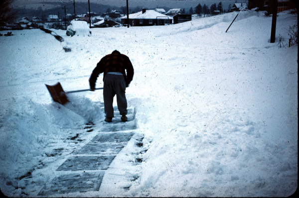 A man in a red plaid jacket holds a shovel and works his way through the heavily collected snow from his yard. Behind him is a path of stepping stones. Ahead is a white landscape with a buried car and homes with their roofs covered in snow.