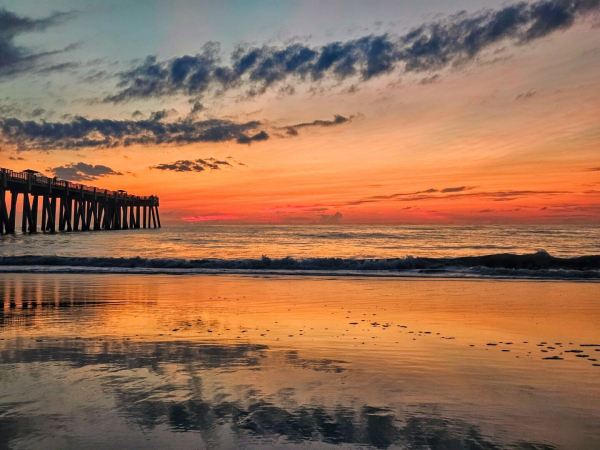 Smokey like clouds hover above layers of color rising from the horizon looking out over the Atlantic Ocean's surf from the wet sands of a beach. The colors stacked in order of intensity with lighter shades of orange on top leading to hot red shades at the horizon.  An immense wooden pier reaches far out into the tides while waves crash, one after another onto the soft wet sand. The horizon's spectrum of colors reflects back from the beach as the water recedes back, exposing bubbles and shells left behind.