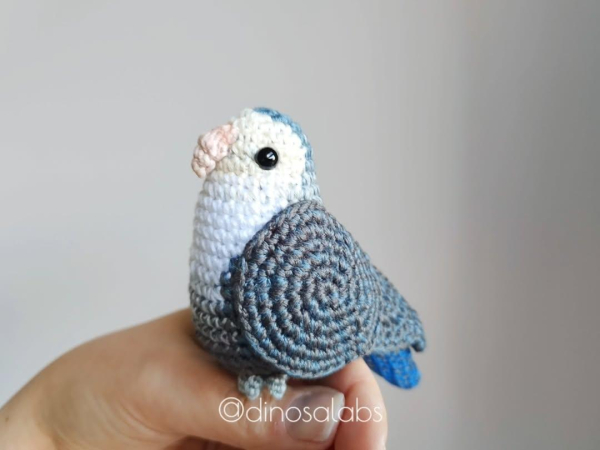 Agapornis tejido a crochet (amigurumi) imitando un pelaje en tono blanco gris y azul.

Se presenta sentadito en una mano como si estuviese posándose