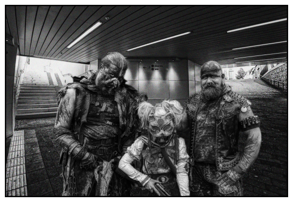 Three apocalyptic world style cosplayers standing facing the camera in an underpass. Behind them we see steps on the left and a ramp on the right leading up to bright daylight. The cosplayers are two burly men like bikers on the right and left and a much smaller masked figure between them, possibly a girl. 