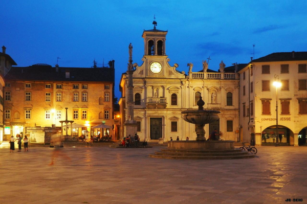 Farbbild - ein Teil der Piazza Matteotti in Udine kurz vor Sonnenuntergang im Sommer. In Bildmitte ein Springbrunnen, links weiter hinten eine Säule. Etwas weiter hinten stattliche historische Gebäude. In der Mitte die Chiesa di San Giacomo aus dem 14. Jahrhundert.