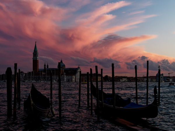 The image captures a stunning sunset over Venice, Italy, with dramatic pink and orange hues painted across the sky. In the foreground, gondolas gently rest on the water, silhouetted against the vibrant backdrop. Wooden mooring posts rise from the lagoon, adding depth and structure to the composition. The iconic church of San Giorgio Maggiore is prominently featured in the midground, its elegant dome and bell tower standing in graceful contrast to the fiery sky. The rippling water reflects the warm tones of the sunset, creating a serene and romantic atmosphere that embodies the timeless charm of Venice.