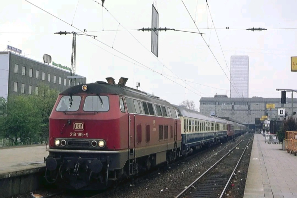 Das Bild zeigt eine Diesellokomotive der Baureihe 218 der Deutschen Bahn mit der Betriebsnummer 218 189-9, die einen klassischen Reisezug mit blau-beigen Wagen durch einen Bahnhof zieht. Die Lok ist in typischem verkehrsrotem Farbton gehalten und hat ein markantes Frontdesign mit zwei großen Scheinwerfern und zwei kleineren Lichtern darunter. Der Bahnhof hat ein langes Bahnsteigdach, Oberleitungen und zahlreiche Schilder, darunter ein gelbes Schild mit einer schwarzen Blitzmarkierung. Im Hintergrund sind moderne Bürogebäude zu sehen, eines davon mit einer Glasfassade, sowie ein größeres Gebäude mit Baugerüst oder Schutzverkleidung. Der Bahnsteig ist belebt mit wartenden Fahrgästen, und der Himmel ist grau, was auf einen bewölkten Tag hinweist.

