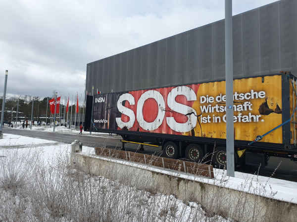 Großer LKW mit der Aufschrift SOS - die deutsche Wirtschaft ist in Gefahr und dem INSM-Logo. Hintergrund ist schwarz-rot-gold. Im Hintergrund sieht man SPD-Fahnen und das Berliner Messegelände, auf dem der SPD-Parteitag stattfand. Es liegt Schnee.