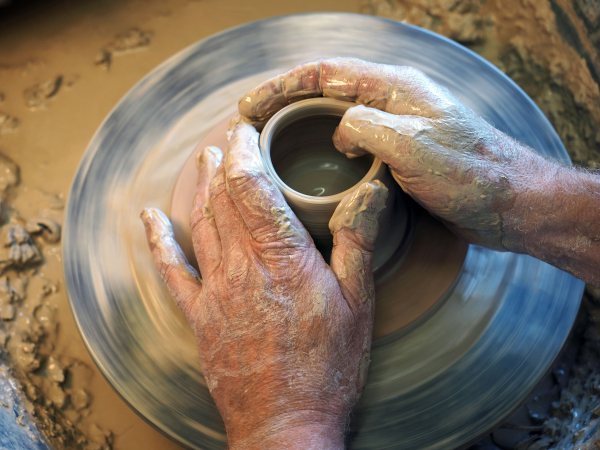 Zwei Hände umfassen einen Tonrohling auf der sich drehenden Töpferscheibe und formen ein Gefäß. Der Fokus der Aufnahme liegt auf den Händen.

Two hands clasp a clay blank on the rotating potter's wheel and form a vessel. The focus of the shot is on the hands.