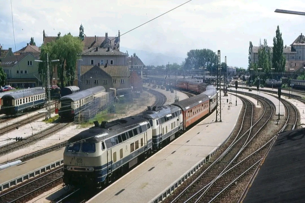 Ein langer Reisezug verlässt einen Bahnhof, gezogen von zwei Diesellokomotiven der Baureihe 218 in beige-blauer Farbgebung. Die Loks fahren in Doppeltraktion und stoßen dunklen Abgasrauch aus. Der erste Wagen des Zuges ist auffällig orange, während die weiteren Wagen eine klassische Schnellzuglackierung in Rot und Beige haben.

Der Bahnhof besteht aus mehreren Gleisen mit Bahnsteigen, und in der linken Bildhälfte stehen abgestellte Reisezugwagen in blauer Farbgebung auf Nebengleisen. Im Hintergrund sind niedrige Häuser mit roten Dächern zu sehen, dahinter erstreckt sich eine hügelige oder bergige Landschaft. Die Szenerie wirkt sommerlich, mit klarem Himmel und grünen Bäumen entlang der Gleise.

