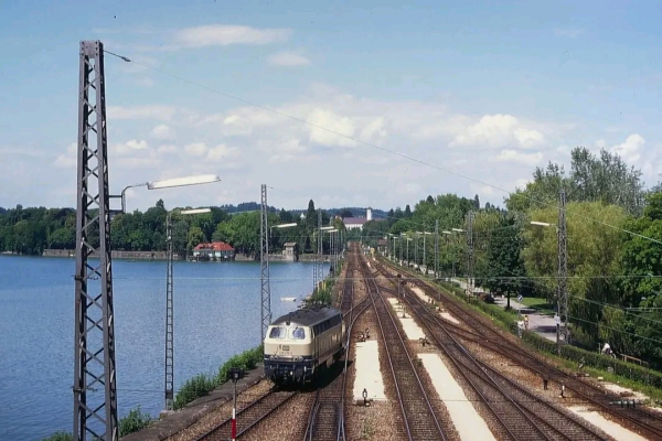 Eine Diesellokomotive der Baureihe 218 in beige-blauer Lackierung fährt auf einem eingleisigen Streckenabschnitt entlang eines Sees. Links des Gleises befindet sich das Wasser, gesäumt von Oberleitungsmasten mit alten Lampen. Im Hintergrund erstreckt sich eine idyllische Landschaft mit dichtem Grün, einem roten Gebäude am Seeufer und einem großen, hellen Bauwerk weiter hinten.

Rechts der Lokomotive verlaufen mehrere parallel geführte Gleise mit Bahnsteigen, auf denen einige Menschen zu sehen sind. Der Himmel ist blau mit wenigen weißen Wolken, die Szene wirkt sommerlich. Die Perspektive zeigt eine weite, gerade Strecke, die sich in der Ferne verliert, und betont die Eisenbahnromantik inmitten der Natur.

