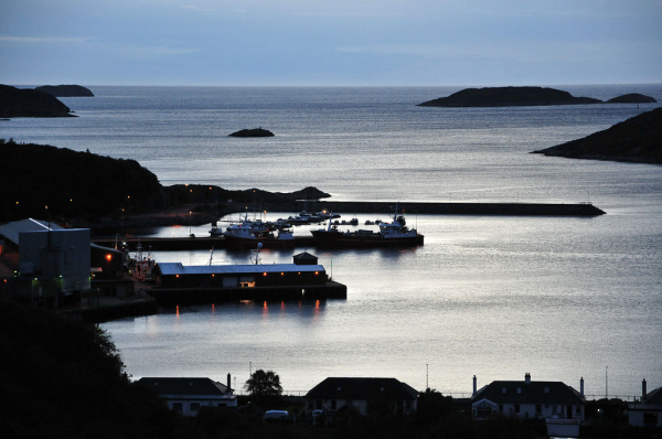 The harbour at Lochinver on the west coast of Sutherland in late evening light. The image shows the harbour from above with the evening light reflecting off it. Silhouettes of houses are in the foreground, and of parts of the harbour on the left. The street lights are on. Dark shapes of islands can be seen out in the loch.