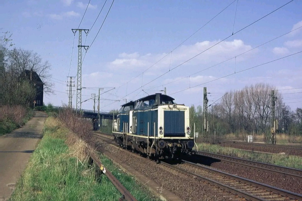 Zwei Diesellokomotiven der Baureihe 212 in beige-blauer Lackierung fahren in Doppeltraktion über eine zweigleisige Bahnstrecke. Die Loks sind aus einer rückwärtigen Perspektive zu sehen, mit ihrem markanten großen Lüftergitter am Führerstand.

Links neben der Strecke verläuft ein leicht ansteigender Weg mit Grasbewuchs, der zu einem alten, dunklen Gebäude führt. Im Hintergrund überspannt eine Brücke die Gleise, und mehrere Oberleitungsmasten ragen in den blauen Himmel mit wenigen weißen Wolken. Rechts der Strecke stehen weitere Masten und Bäume mit noch spärlichem Laub, was auf Frühling oder frühen Herbst hindeutet.

