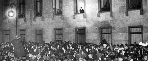 A historical black and white photo showing a large crowd gathered at night in front of a building, with people visible in the building's windows, suggesting a significant public event or announcement.