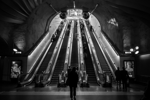 Black and white photo of escalators at a subway station. In the foreground a dark sihouette of a person walking towards the escalators.