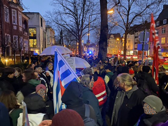 Menschen demonstrieren auf einer Straße. Einige tragen Fahnen. Zwei weiße Regenschirme und Demoplakate der Omas gegen Rechts sind deutlich zu erkennen.