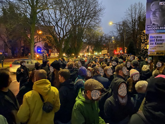 Vielen Menschen auf einer Straßenseite. Einige wenige Demoschilder sind zu sehen. Am rechten Rand ein CDU Wahlkampfplakat mit dem Kandidaten Mathias Middelberg.