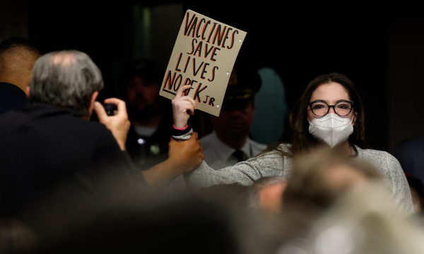 Photo of a woman in a grey shirt, white respirator and glasses holding a sign that says “vaccines save lives no RFK Jr”