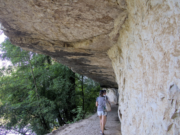 2 personnes de dos, l'une derrière l'autre, en short + t-shirt et un sac à dos marchent sur un chemin creusé dans la pierre du côté droit. Le chemin est également surplombé par la roche, à gauche on voit des arbres le long d'une rivière qu'on distingue en-dessous.