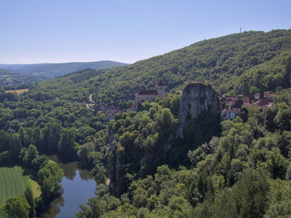 Photo de paysage vallonné avec beaucoup de forêt, prise en hauteur. On voit une large rivière en contrebas bordée d'arbres, une culture de maïs sur la berge à gauche. À droite, un rocher massif à forme arrondie en haut masque partiellement un village dont on voit l'église et plusieurs maisons de part et d'autre. Le ciel est bleu sans nuage.