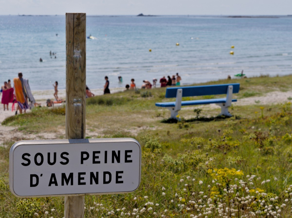 Au 1er plan, un panneau incomplet : le haut du poteau est vide, et le panneau dessous indique "Sous peine d'amende". Au second plan, une dune partiellement végétale, on voit un banc bleu et blanc qui fait face à la mer. Au 3e plan, des gens sur une plage, et en arrière plan la mer (et des baigneurs) sous le soleil, le ciel est sans nuage. Au fond on aperçoit des îlots sombres.