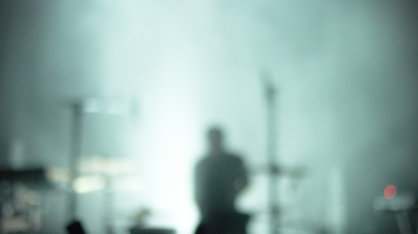 Photo de concert floue : on distingue une silhouette et des cymbales, pieds de micros… l'ambiance générale est vert-bleu pâle avec une lumière plus blanche au centre, derrière la silhouette.