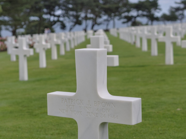 Cimetière militaire en Normandie, des alignements de croix blanches dans l'herbe verte, au 1er plan une croix où est écrit : Patrick J. Brown 
SGT 8 INF 4 DIV
New York June 24 1944
En arrière plan quelques arbres devant la mer et le ciel.