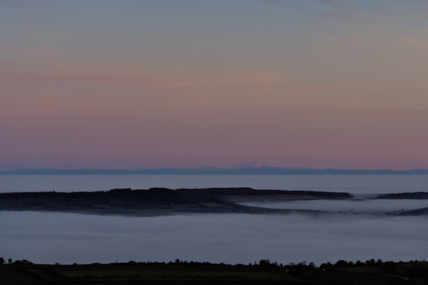 Photo prise au coucher de soleil en hiver, en hauteur depuis un mont : au 1er et second plan, on voit des crêtes sombres émerger d'une mer de brouillard qui se poursuit loin. En arrière plan, d'autres reliefs, dont une montagne enneigée éclairée par une lueur rose sur la droite. Le ciel est en dégradé de rose-orange-bleu.